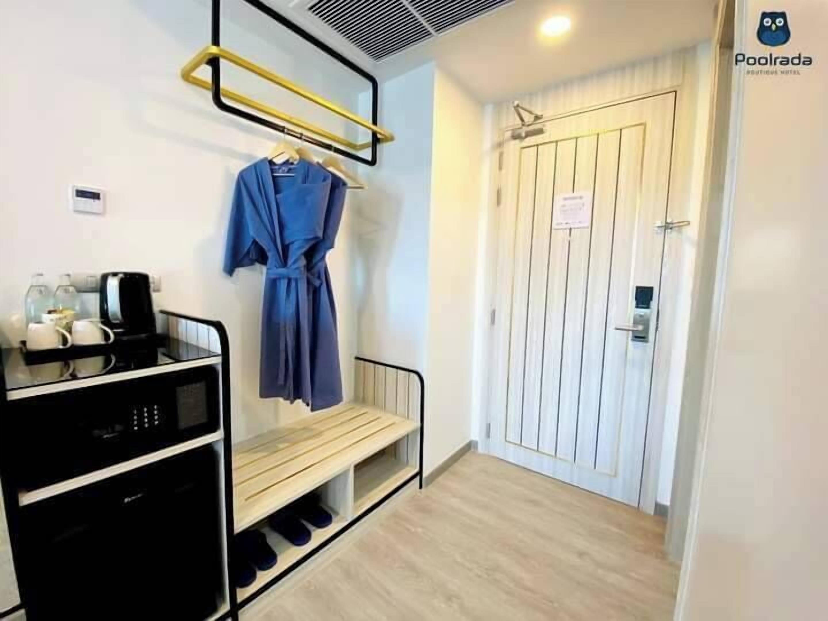 Habitación de hotel con kitchenette, bata colgada en un perchero, banco de madera y una puerta con un cartel.