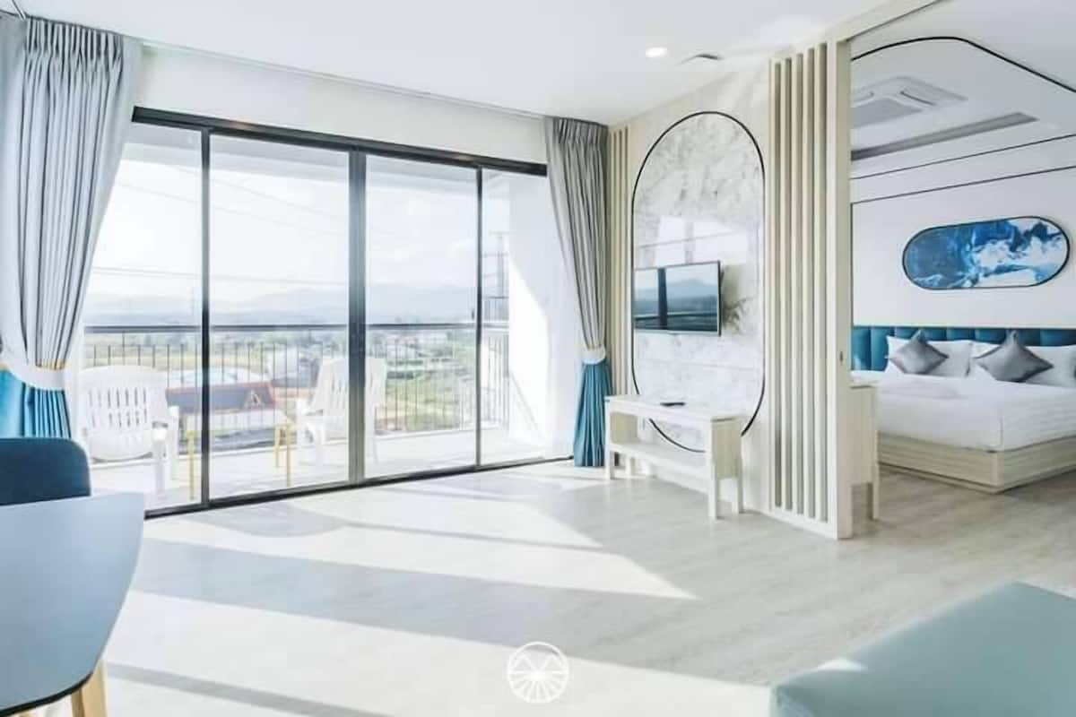 A modern hotel room with a large balcony, a bed with a blue headboard, a desk, and a TV mounted on the wall.