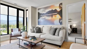 Een moderne woonkamer met een groot raam, een bank, een salontafel en een schilderij van een berglandschap.