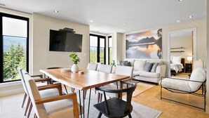 Een moderne woonkamer met een houten eettafel, witte banken en een groot schilderij van een meer.