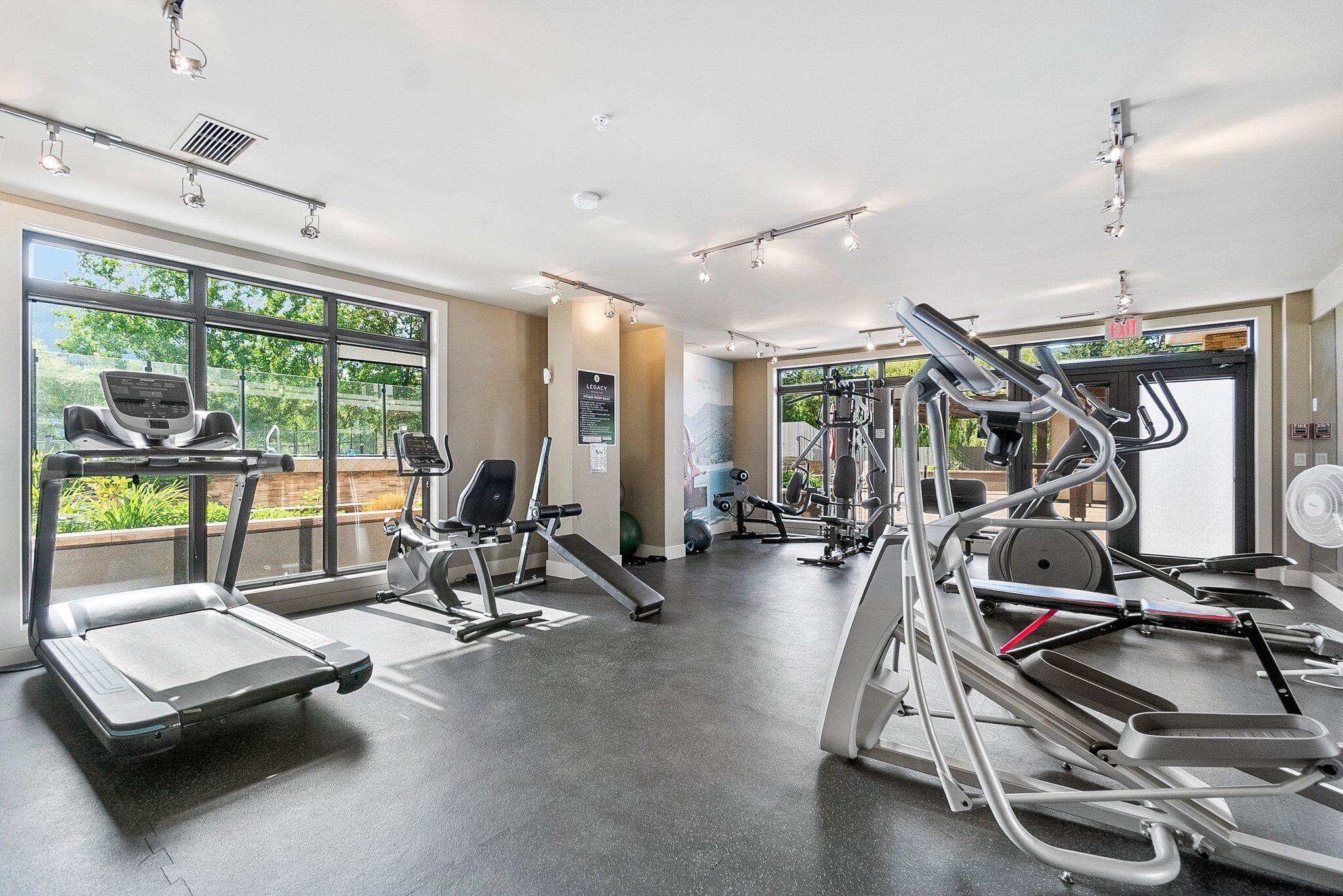 A modern gym with treadmills, ellipticals, and weight machines. Large windows provide natural light and views of greenery outside.