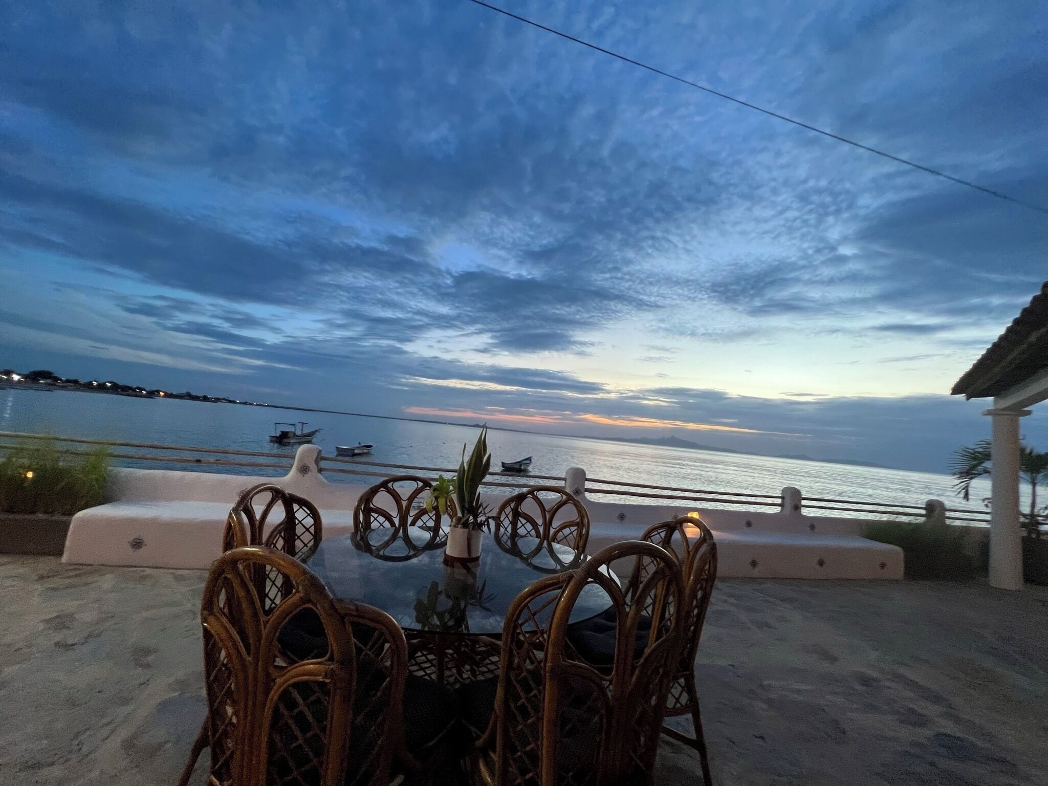 An outdoor seating area with wicker chairs and a table, overlooking a calm sea with boats and a sunset sky.
