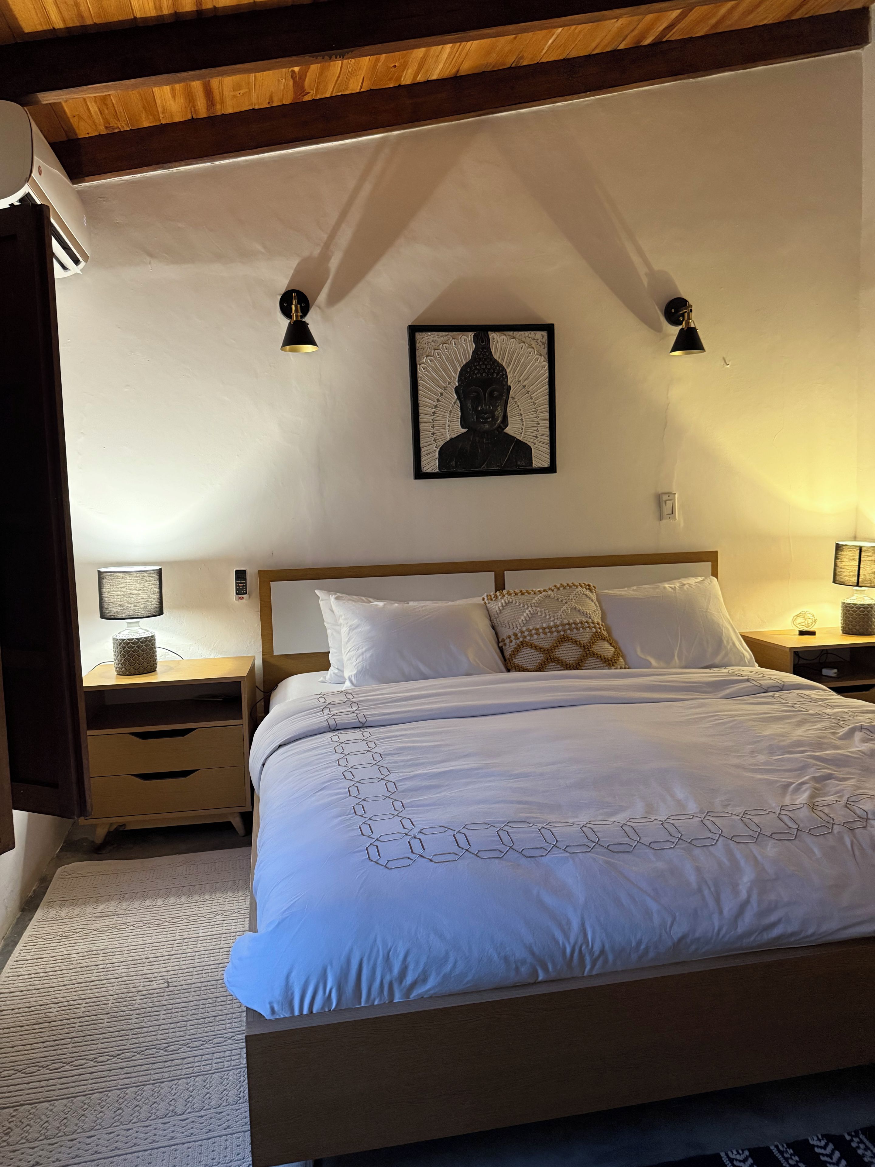 Une chambre avec un lit en bois, du linge de lit blanc, une statue de Bouddha fixée au mur, des lampes de chevet et une table de chevet avec une lampe.