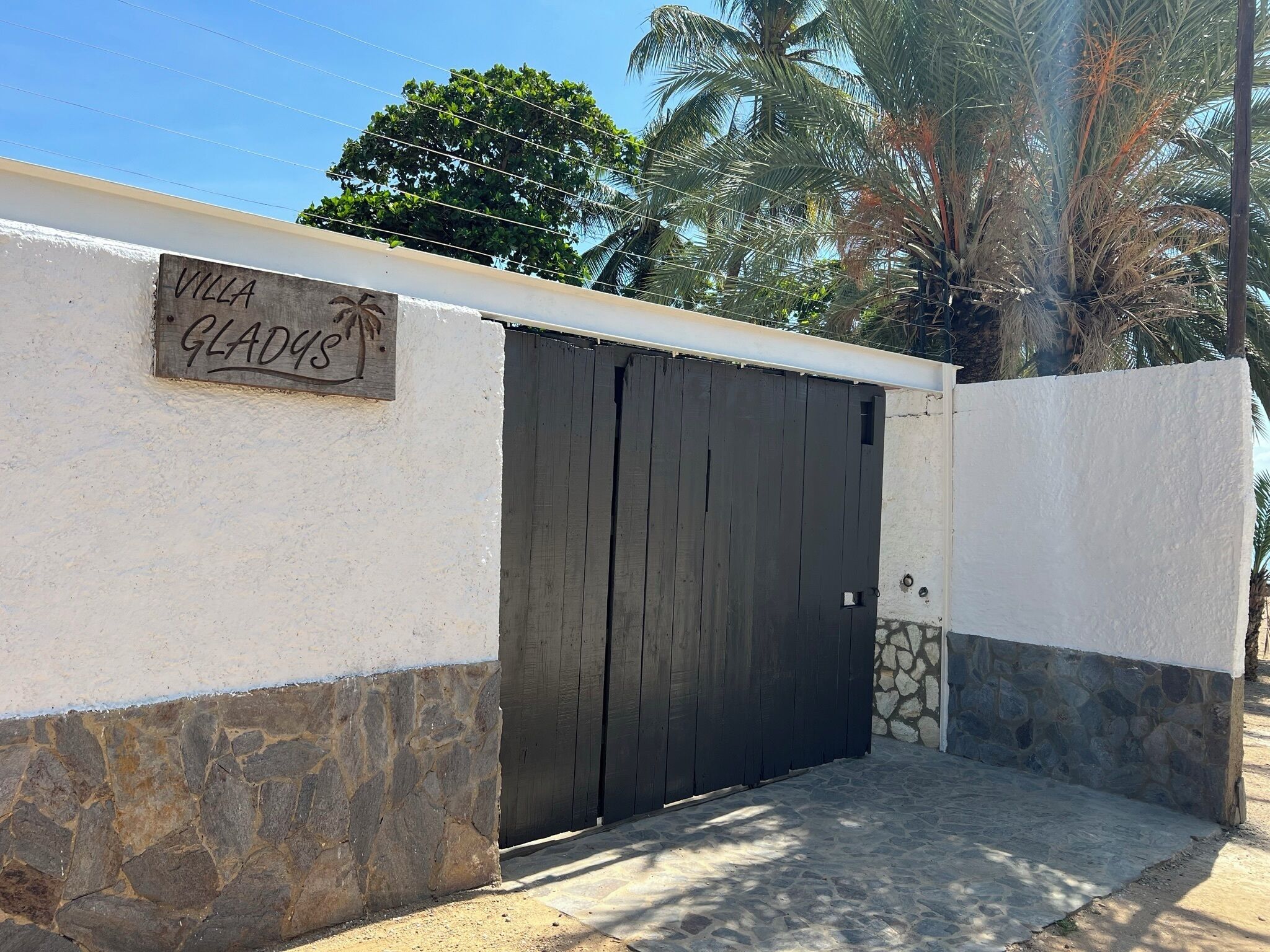 En villa kaldet "Villa Gladys" med en mørk træport, stenvægge og et palmetræ i baggrunden.