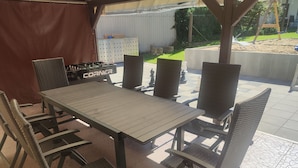 Une terrasse extérieure avec une table et des chaises, un espace abrité et une vue sur un jardin avec un portique.