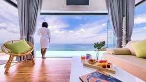 Ein Zimmer mit großem Fenster zum Meer, ein Korbstuhl, eine Couch und ein Frühstücksbrettchen.