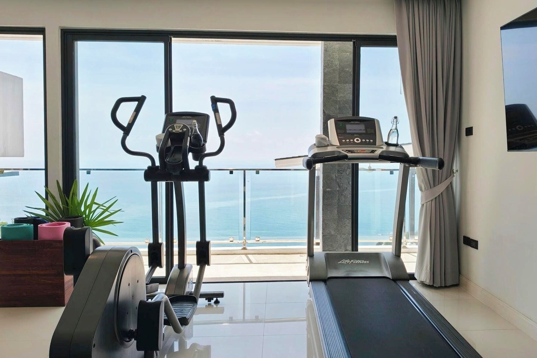 A home gym with a treadmill and elliptical machine, large windows with a view of the sea, and a potted plant.