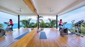 Ein geräumiges Fitnessstudio mit Holzboden, Trainingsgeräten und großen Fenstern mit Blick auf den Ozean.