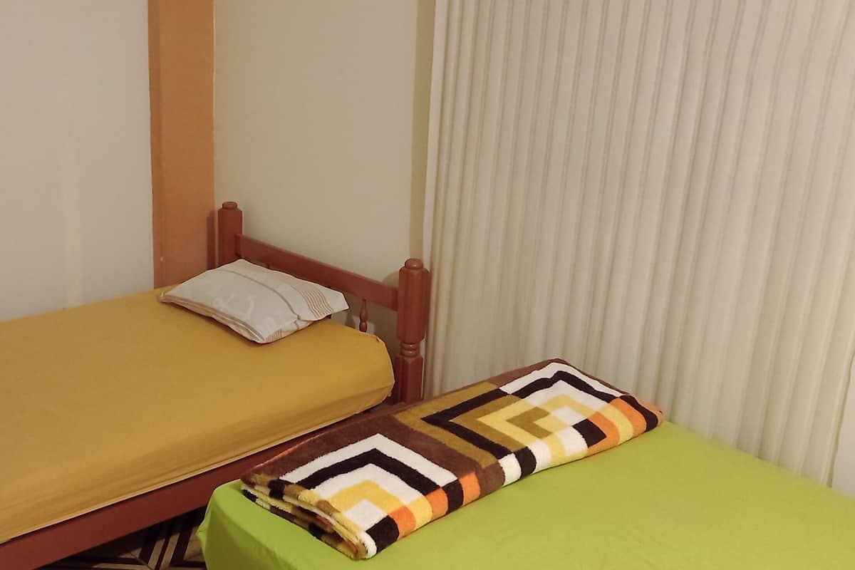 Ein kleines Zimmer mit einem Einzelbett, einem grünen Tisch und einem Fenster mit Vorhängen.