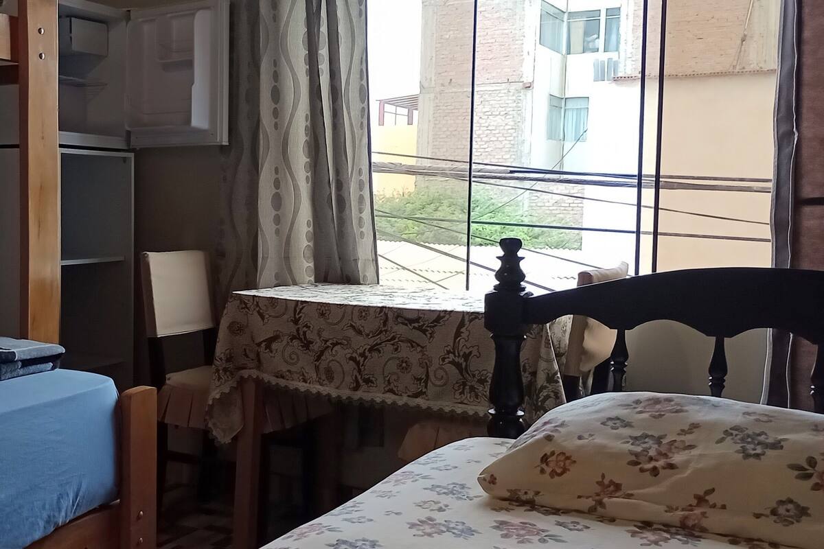 Ein Zimmer mit Etagenbetten, einem kleinen Tisch mit Blumenmuster-Tischdecke, einem Stuhl und einem Fenster mit Vorhängen.