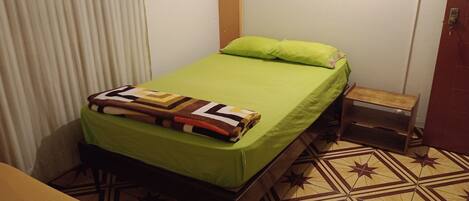 Una cama individual con cubrecama verde y manta estampada, una mesita de noche de madera, un pequeño banco de madera y un suelo con estampado.
