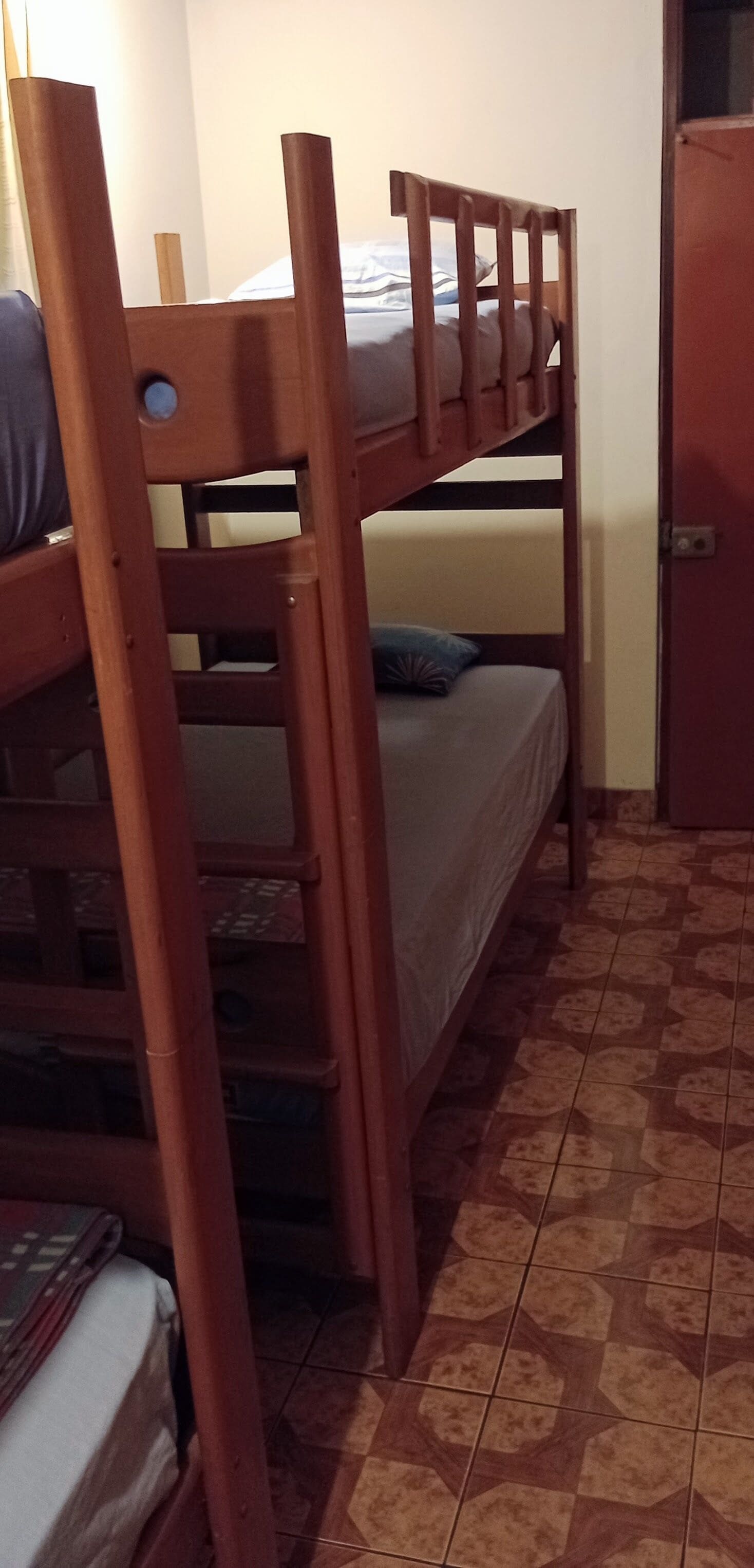 A bunk bed with a wooden frame, a lower bunk with a mattress and pillow, and a lower bed with a mattress and pillow. There is a door to the right and a wall with a light source.