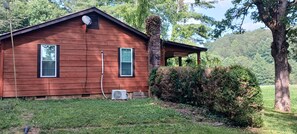 A wooden cabin with two windows, an air conditioning unit, and a covered porch.