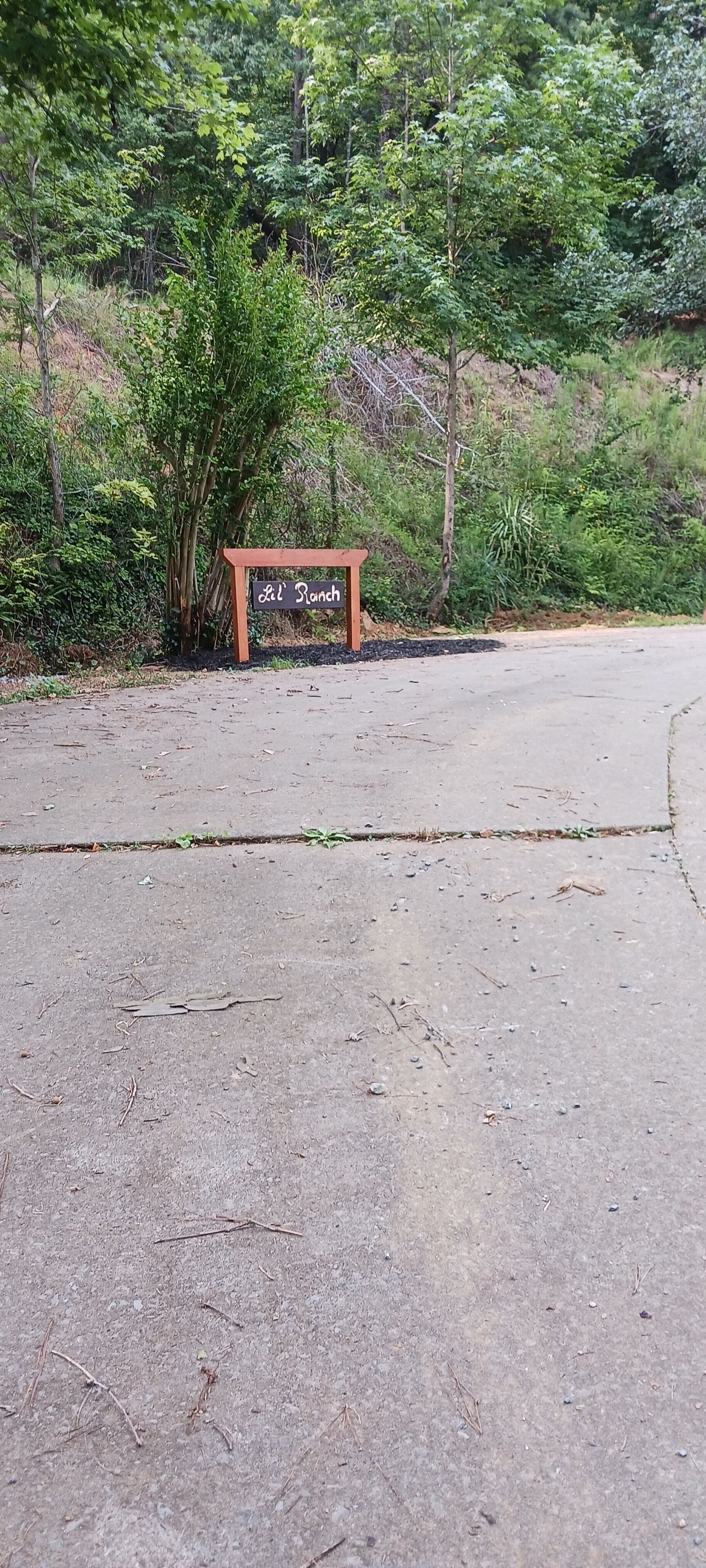 A sign reading "Lil Ranch" in a natural setting with trees and a dirt path.