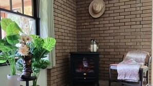 Een kamer met een bakstenen muur, een houten vloer, een raam met gordijnen, een plant in een pot, een hoed aan de muur, een stoel met een deken en een kleine tafel met een vaas.