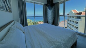 Une chambre d’hôtel avec un grand lit, deux fenêtres avec des rideaux, une vue sur la mer et, au loin, un bâtiment avec un toit rouge.