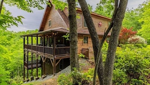 A wooden cabin with a porch and a deck, surrounded by trees and greenery. - Cabin log home w view, hot tub, game room. Community pool 4 bedroom Sleeps 10-12 (Deep Gap)