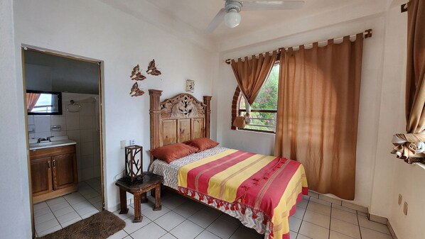 A bedroom with a bed, a wooden headboard, a window with curtains, a bedside table, and a mirror. - Sebas (Yelapa)