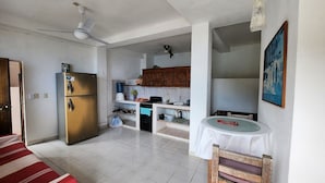 Eine kompakte Küche mit Kühlschrank, Herd und Spüle. Ein Esstisch mit Stühlen und ein Deckenventilator sind vorhanden.