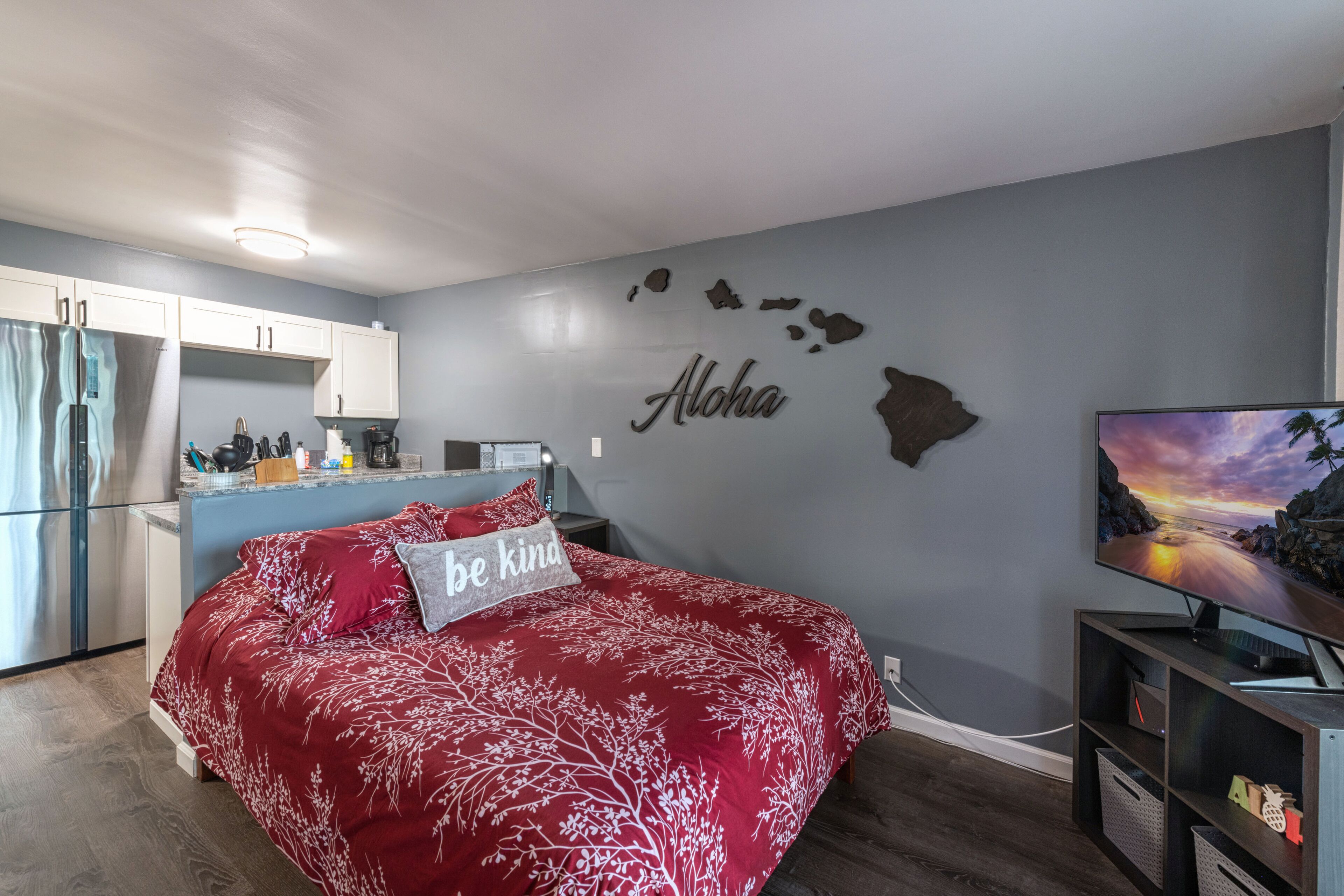 A bedroom with a bed, a TV, a kitchen area, and a wall with a map and the word "Aloha".