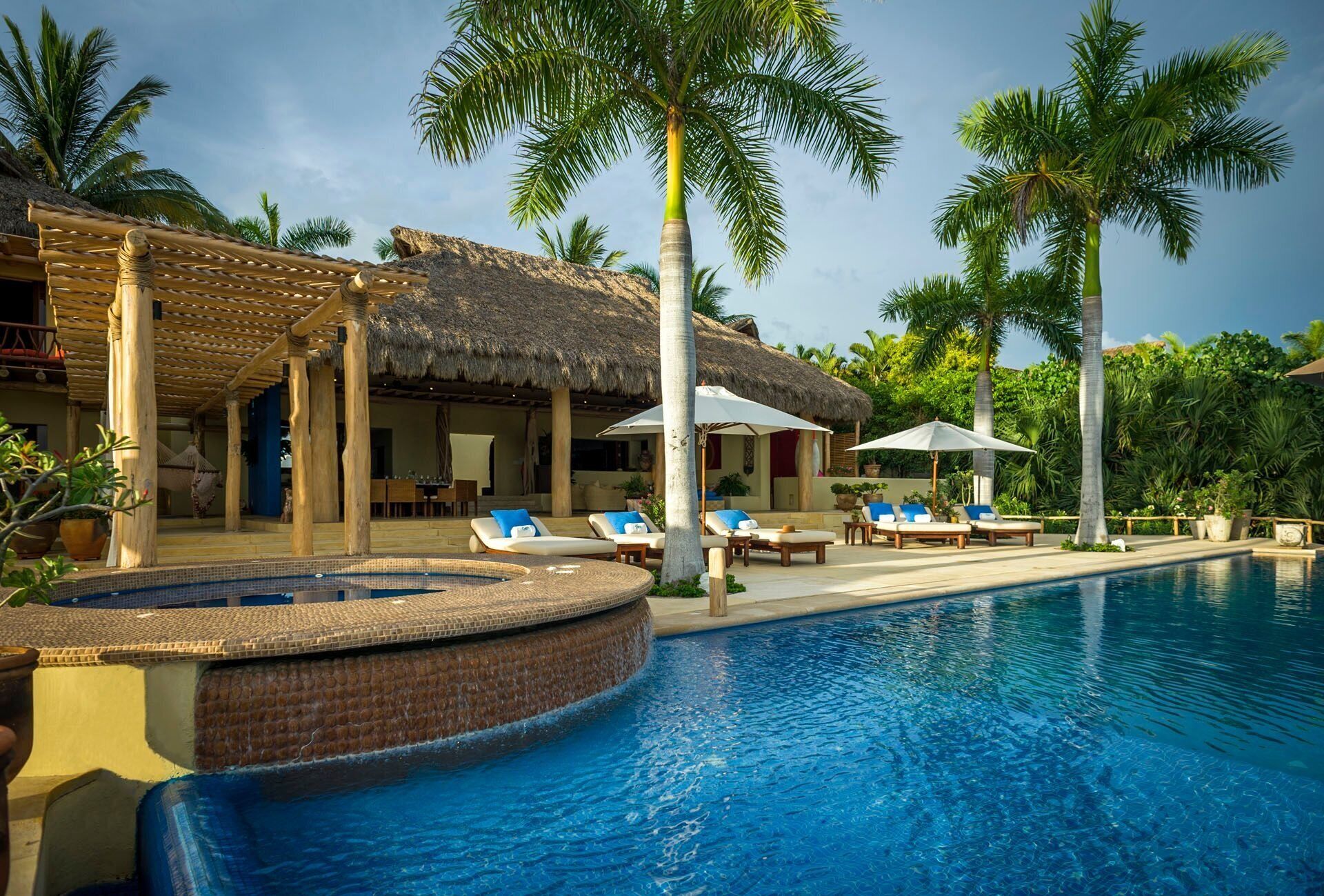 A poolside area with lounge chairs, a hot tub, and a thatched-roof structure.