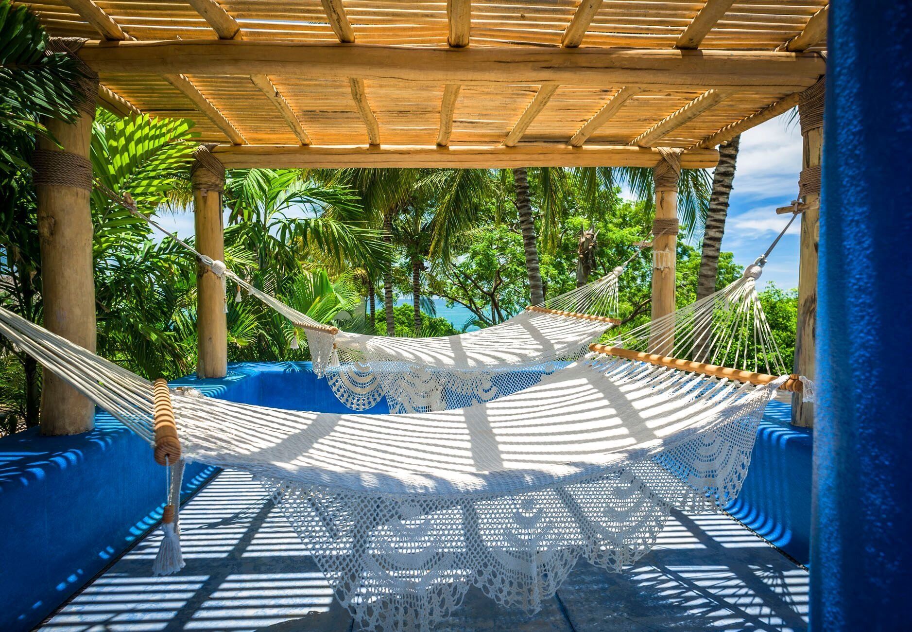 Riippumatto puisen pergola-rakenteen alla, josta avautuu näkymä palmuille ja vesistölle.