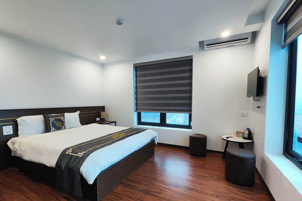 A modern hotel room with a large bed, a bedside table, a small stool, a window with blinds, an air conditioning unit, and a TV mounted on the wall.