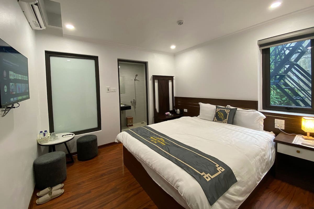 A hotel room with a bed, a bedside table, a TV, a mirror, a window, and a shower area.