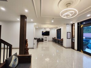 A modern hotel lobby with a reception desk, a TV mounted on the wall, a dining area with chairs, and a large window offering a view of the outdoors. - Apricot Garden Hotel (Sa Pa)