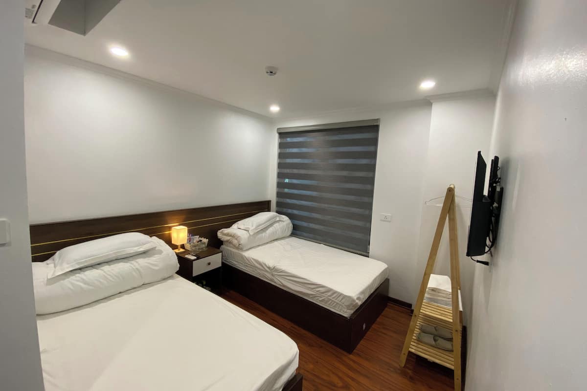 A hotel room with two single beds, a wooden floor, a window with blinds, a bedside table with a lamp, and a wall-mounted TV.
