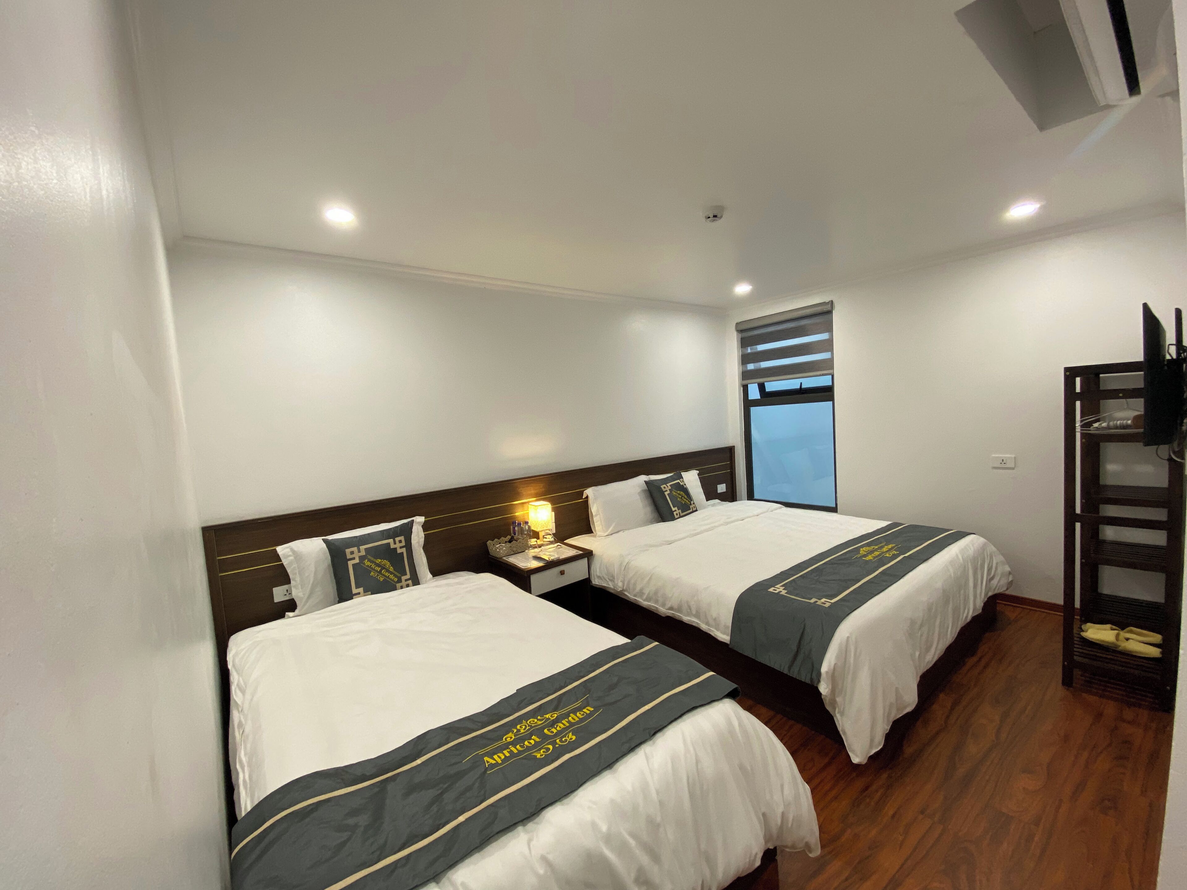 Une chambre d’hôtel avec deux lits, une table de chevet avec une lampe, un téléviseur posé sur un meuble et une fenêtre avec des stores.