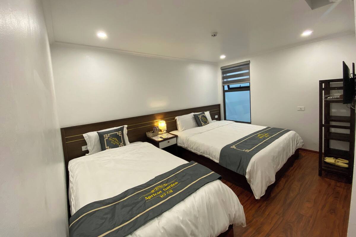 A hotel room with two beds, a nightstand with a lamp, a TV on a stand, and a window with blinds.