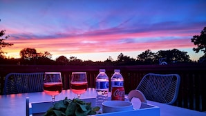 Mesa posta com copos de vinho, garrafas de água e uma garrafa de Coca-Cola em uma varanda com vista para o pôr do sol.