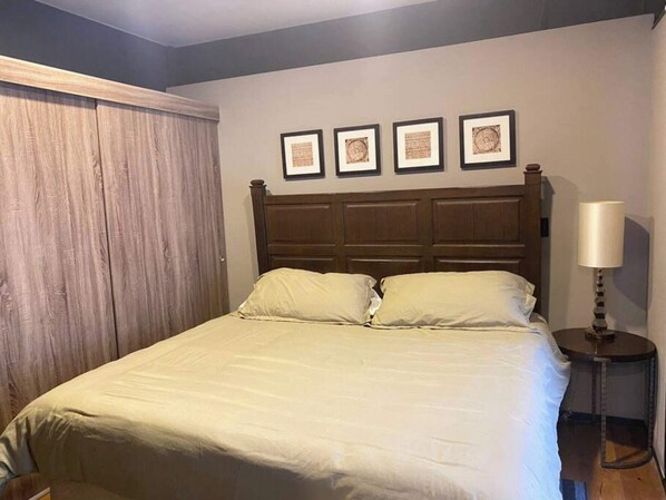 A bedroom with a wooden headboard, a bed with white bedding, a nightstand with a lamp, and three framed pictures on the wall.