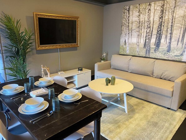 A dining area with a table set for four, a sofa, a flat-screen TV, and a large forest-themed painting.