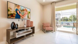 Sala de estar con televisor de pantalla plana, sofá, sillón y vista a una piscina a través de amplios ventanales.