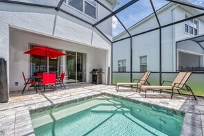 Une piscine entourée de parois en verre, des meubles d’extérieur rouges et un barbecue.
