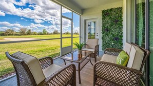 A covered patio with wicker furniture, a glass table, and a view of a grassy field through large windows. - Chic Golf-View Condo - Simply Amazing (Four Corners)