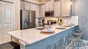 A modern kitchen with white cabinets, stainless steel appliances, a central island with seating, and a hanging light fixture. - Chic Golf-View Condo - Simply Amazing (Four Corners)