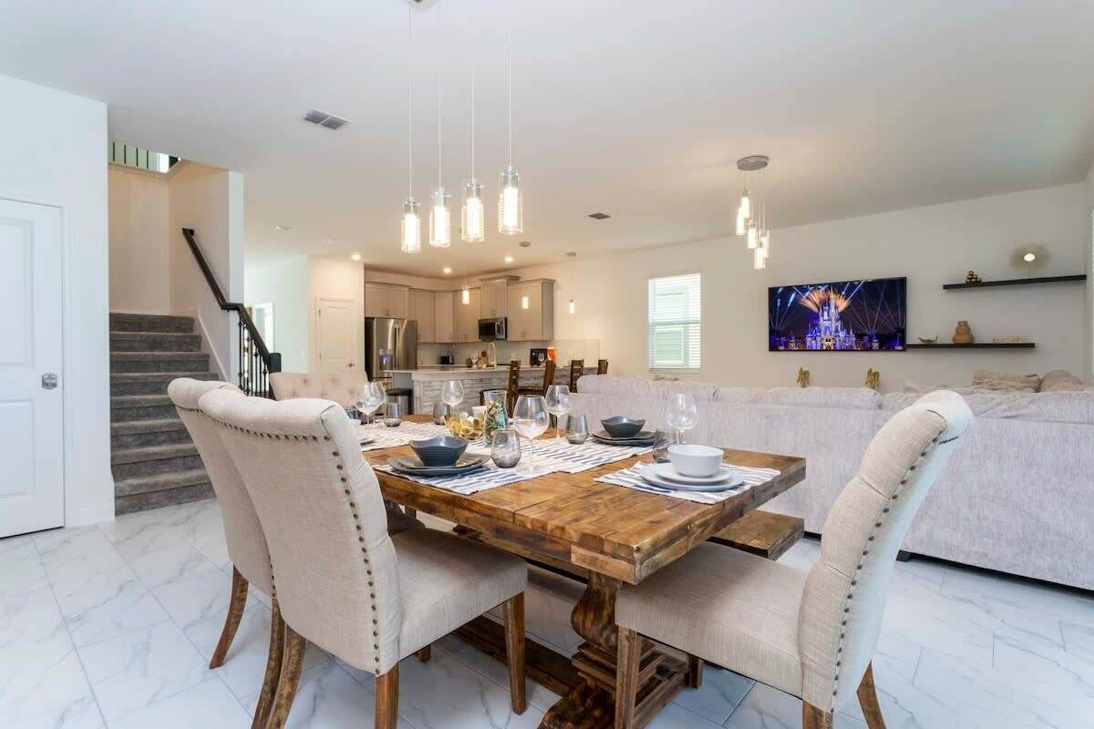 Un espace repas avec une table en bois dressée pour quatre personnes, des chaises à revêtement blanc et des luminaires suspendus.