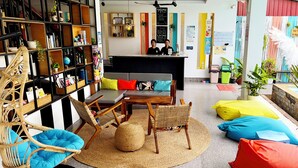 A cozy lounge area with a sofa, chairs, and a small table. There are shelves with books and decorative items, and a reception desk with a sign. - LOTUS SERENITY Hostel (Siem Reap)