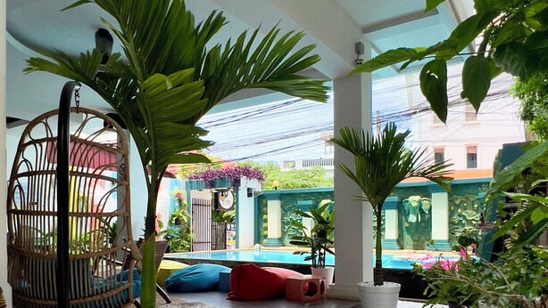 A spacious indoor area with a white pebble floor, surrounded by potted plants and wicker furniture. - LOTUS SERENITY Hostel (Siem Reap)