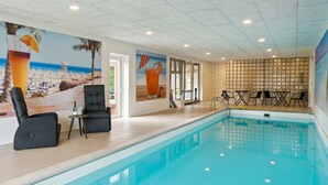 Una piscina interior con un mural con temática playera, un área de descanso con sillas y una mesa, y vistas al exterior a través de un ventanal.