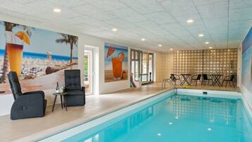 En indendørs swimmingpool med et mural i strandtema, et loungeområde med stole og et bord, samt udsigt til det fri gennem en glasdør.