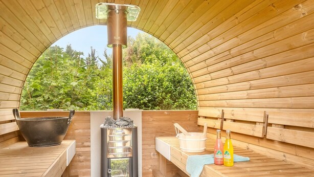 Una sauna con interior de madera, un calentador central, un cuenco negro, un balde de madera y dos botellas de productos de baño.