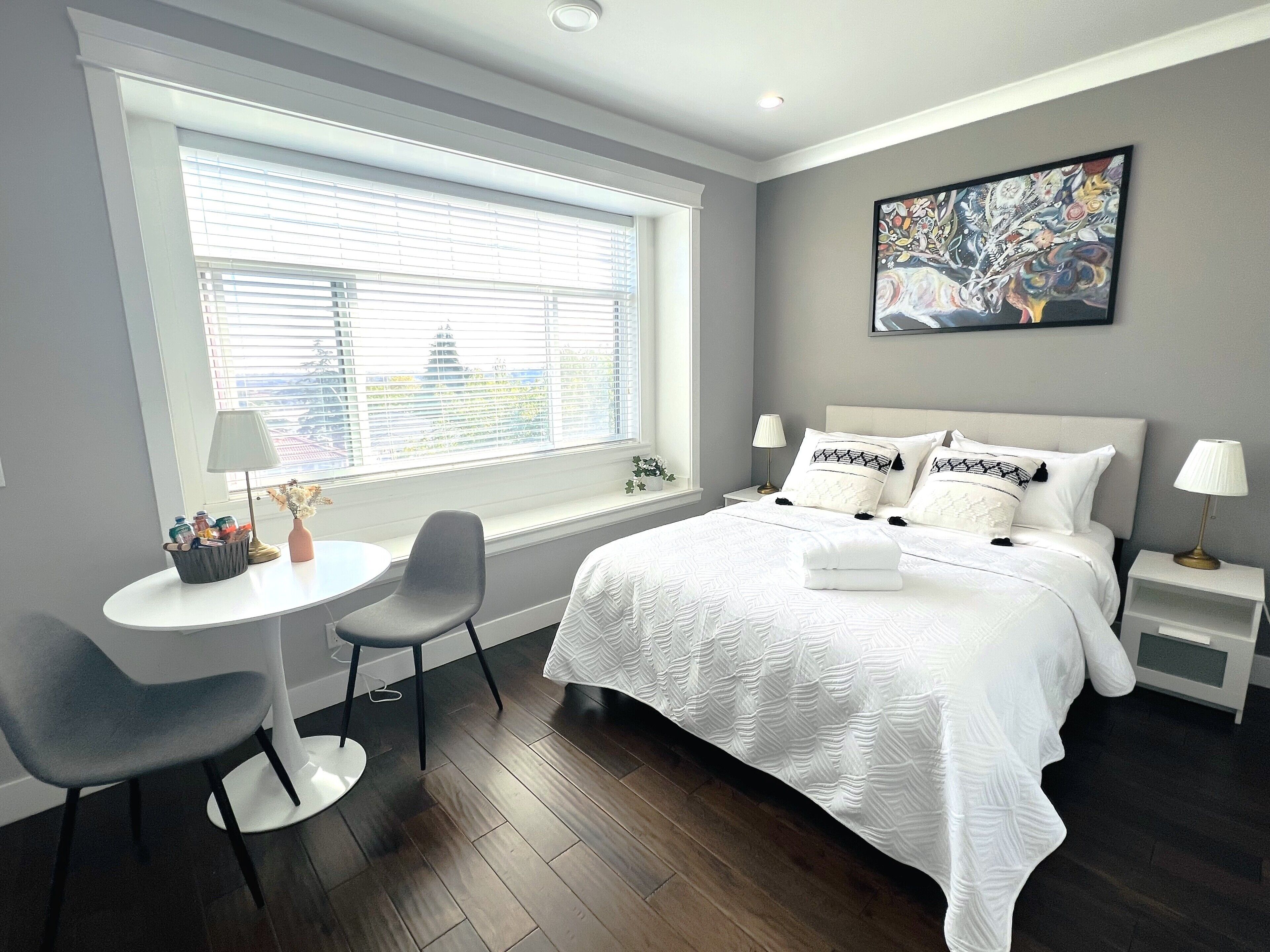 Une chambre moderne avec un grand lit, deux tables de chevet, une petite table ronde et une chaise. Il y a une fenêtre avec des stores, un tableau au mur et une vue sur un bâtiment à l’extérieur.