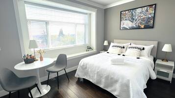 Une chambre moderne avec un grand lit, deux tables de chevet, une petite table ronde et une chaise. Il y a une fenêtre avec des stores, un tableau au mur et une vue sur un bâtiment à l’extérieur.