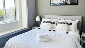 A neatly made bed with white bedding and decorative pillows, a bedside table with a lamp, and a colorful abstract painting on the wall. - Jennifers House (Vancouver)