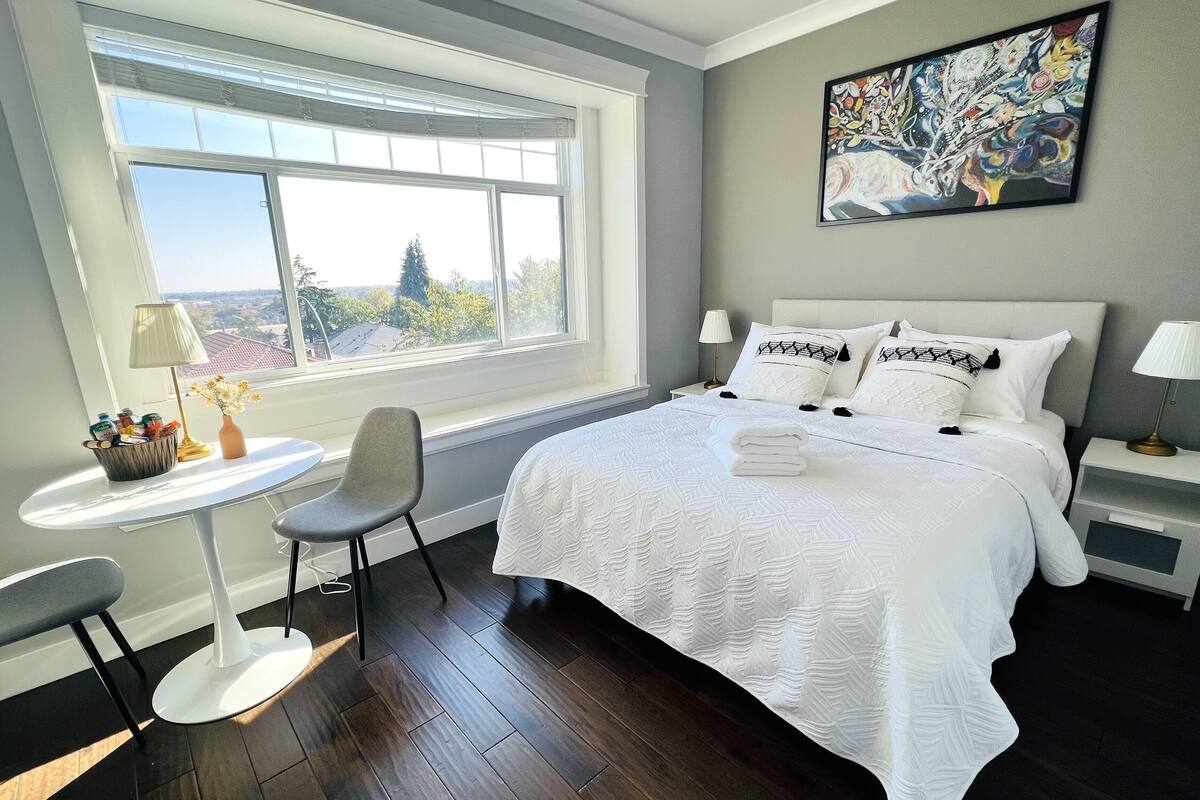 A hotel room with a large bed, a small table with a lamp, a chair, and a view of the outside through a large window.