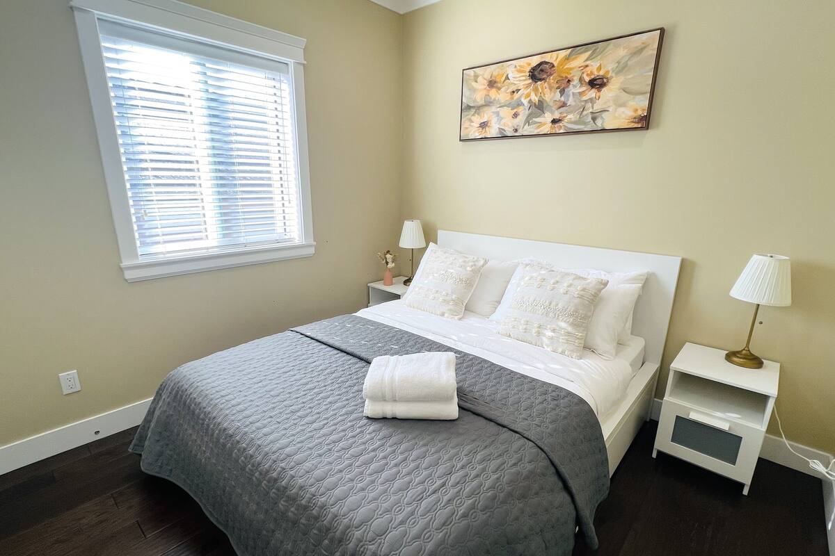 A bedroom with a large bed, a window with blinds, a nightstand with a lamp, and a painting on the wall.